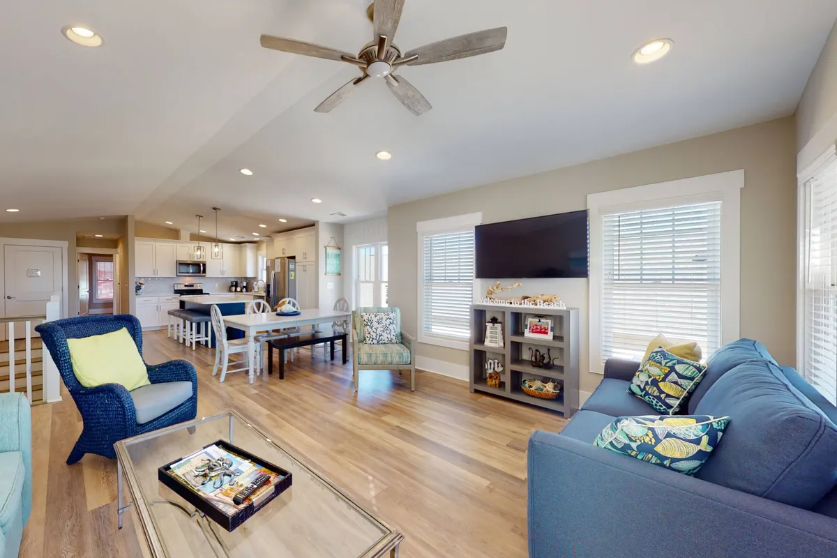 The image shows a welcoming interior with a blue sofa adorned with colorful cushions, a wooden dining table ideal for group meals or board games, and a decorative shelf. The space is airy and bright, with a ceiling fan overhead and clear sightlines between the living and dining areas.