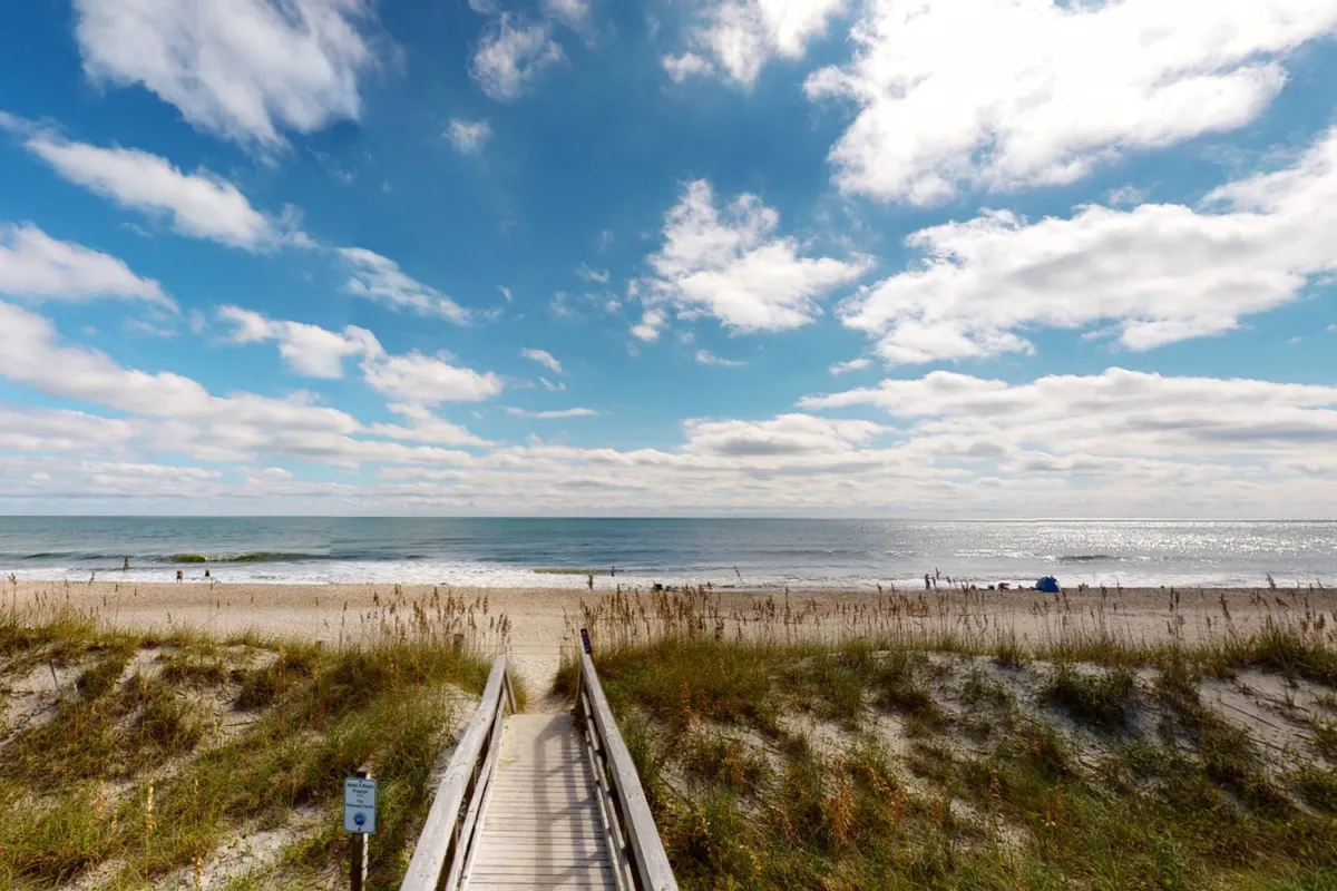 A wooden boardwalk leads through beach dunes to a wide, sandy shoreline with the ocean and blue sky beyond. Children and families can be imagined building sandcastles and collecting seashells, echoing the playful, welcoming mood described in the section.