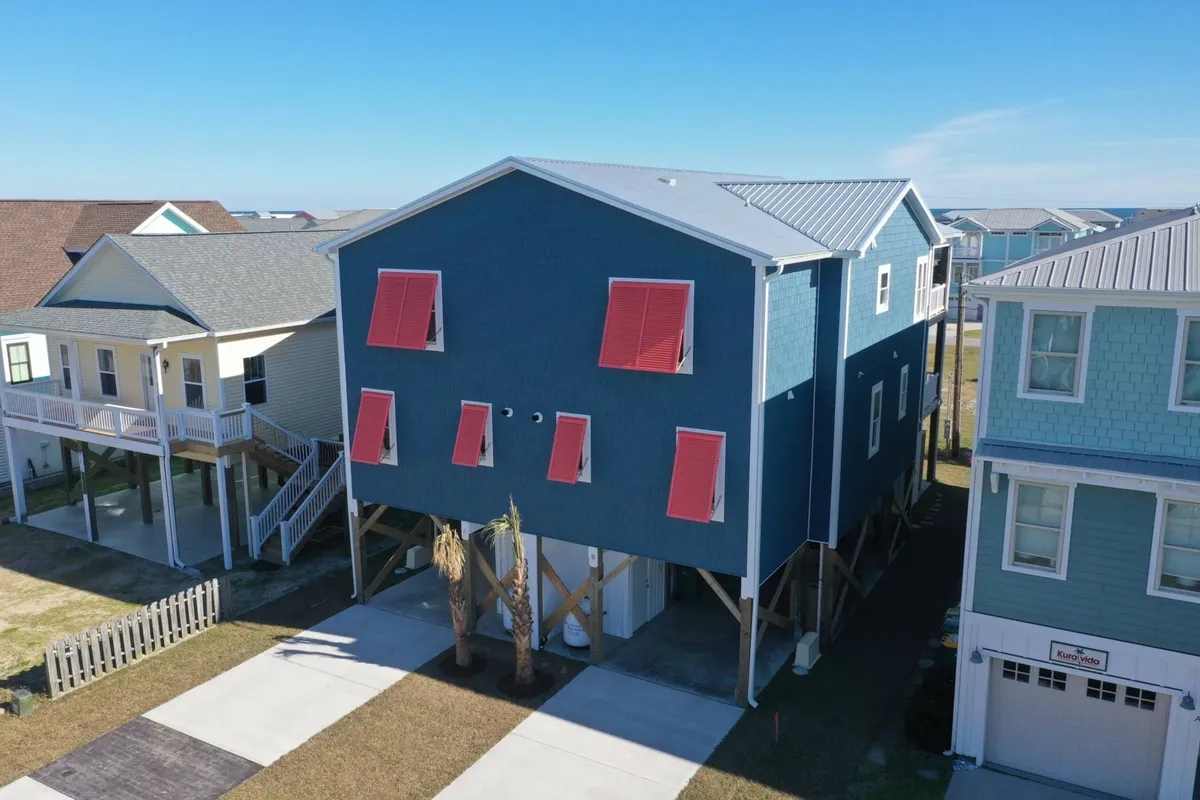 The cheerful exterior of a blue house with red shutters, framed by palm trees and neighboring homes. The porch and yard suggest spaces for family gatherings and casual outdoor enjoyment in a peaceful setting.