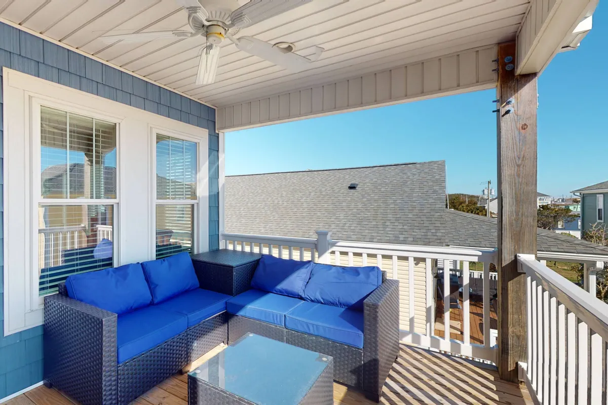 A welcoming outdoor deck furnished with blue cushioned seating, a glass coffee table, and shaded by a ceiling fan. The wooden railing frames tranquil neighborhood views, inviting relaxation and socializing. The deck appears elevated, suggesting views and privacy.