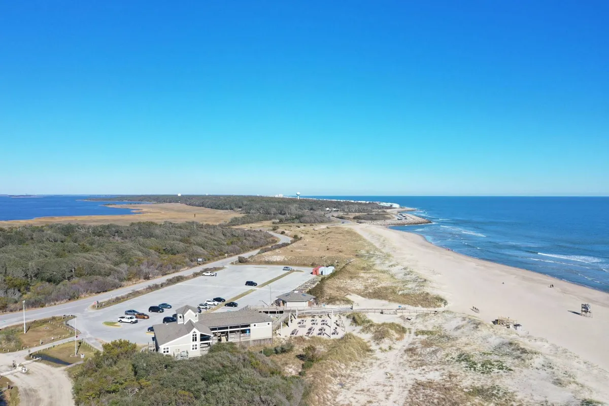 The image features a peaceful beachfront scene with ocean waves washing ashore, a coastal restaurant visible nearby, and a relaxed parking area. The inviting atmosphere suggests opportunities for oceanfront dining and pleasant strolls to local eateries.