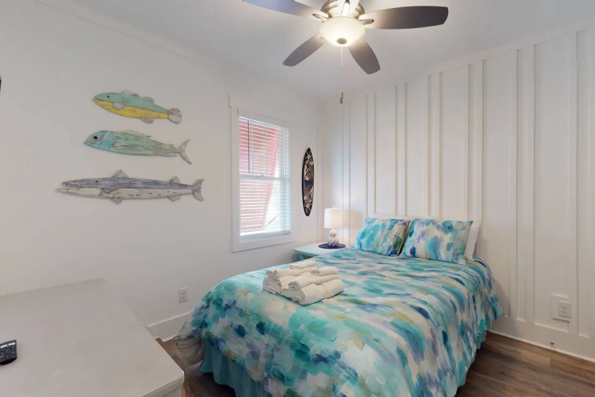 A cozy bedroom with a bed dressed in colorful bedding, decorative fish wall art, and a warm table lamp. Natural light filters in, creating an inviting, restful space ideal for couples or friends.