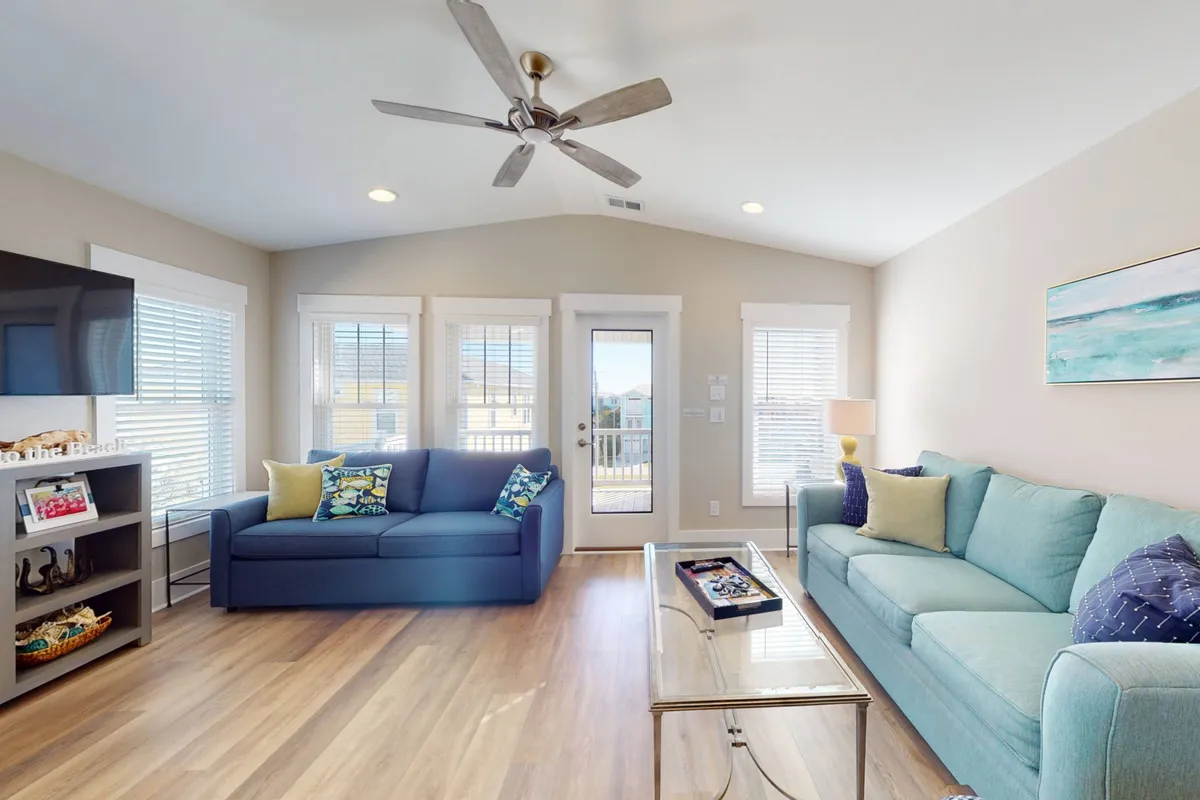 This image features a bright, sun-drenched living room with expansive windows, blue and teal sofas, a glass coffee table, and a ceiling fan. The abundance of natural light and comfortable seating make it ideal for socializing or unwinding. There are visible exterior views through the windows, hinting at the coastal setting.