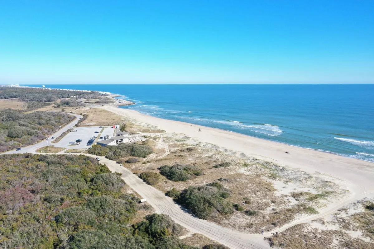 This exterior image captures a tranquil stretch of sandy beach with the ocean gently rolling in, bordered by natural dunes and a nearby parking area. The soft morning or evening light creates a serene coastal ambiance, inviting visitors to imagine peaceful walks along the shore or early morning sunrises.