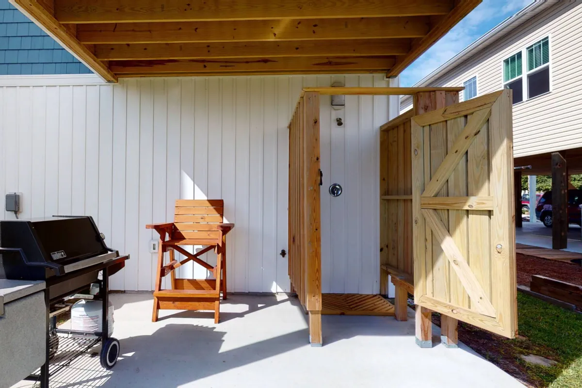 A relaxed outdoor area featuring an outdoor shower, BBQ grill, and a wooden chair set against a backdrop of sunlit siding. The space is casual, practical, and clearly designed for post-beach refreshment and social outdoor dining.