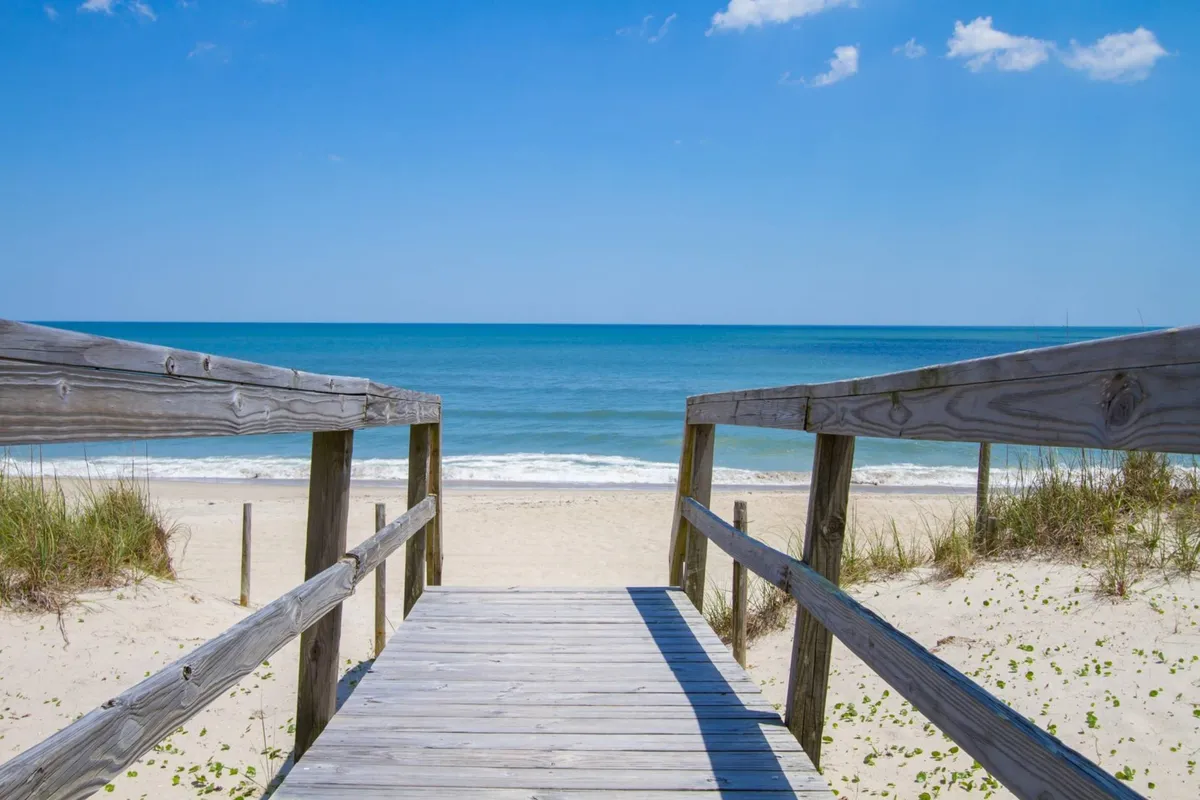 An inviting wooden walkway leads directly to an open view of the ocean and sky, with the sandy beach stretching out ahead. The scene is tranquil, evoking the pleasure of sunset chats or early morning strolls, and embodies the relaxing connection between the home and the outdoors.